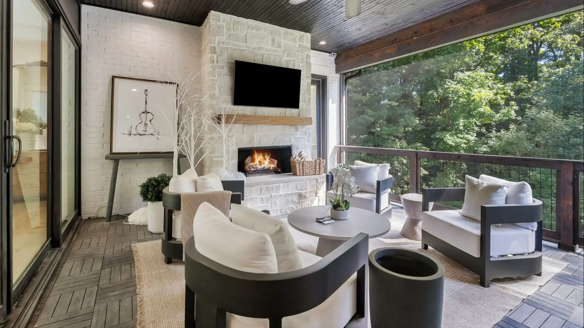 Screened outdoor living space in a Nashville luxury home featuring a stone fireplace, mounted TV, dark wood ceiling, and modern white outdoor furniture overlooking mature trees